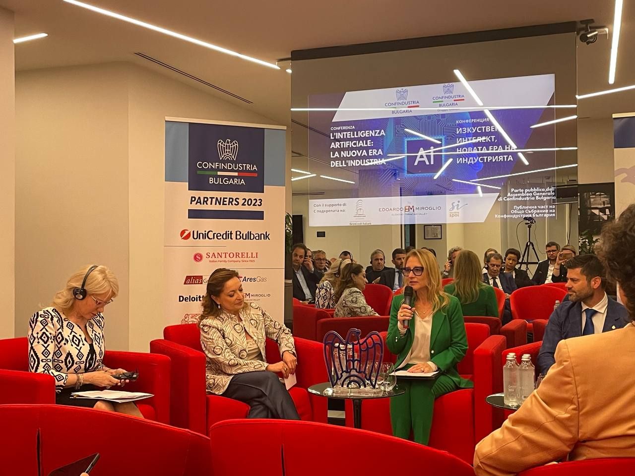 A speaker in a green suit addresses the audience from a red armchair at the Confindustria Bulgaria conference, with the screen behind her reading "L'Intelligenza Artificiale: la nuova era dell'industria"