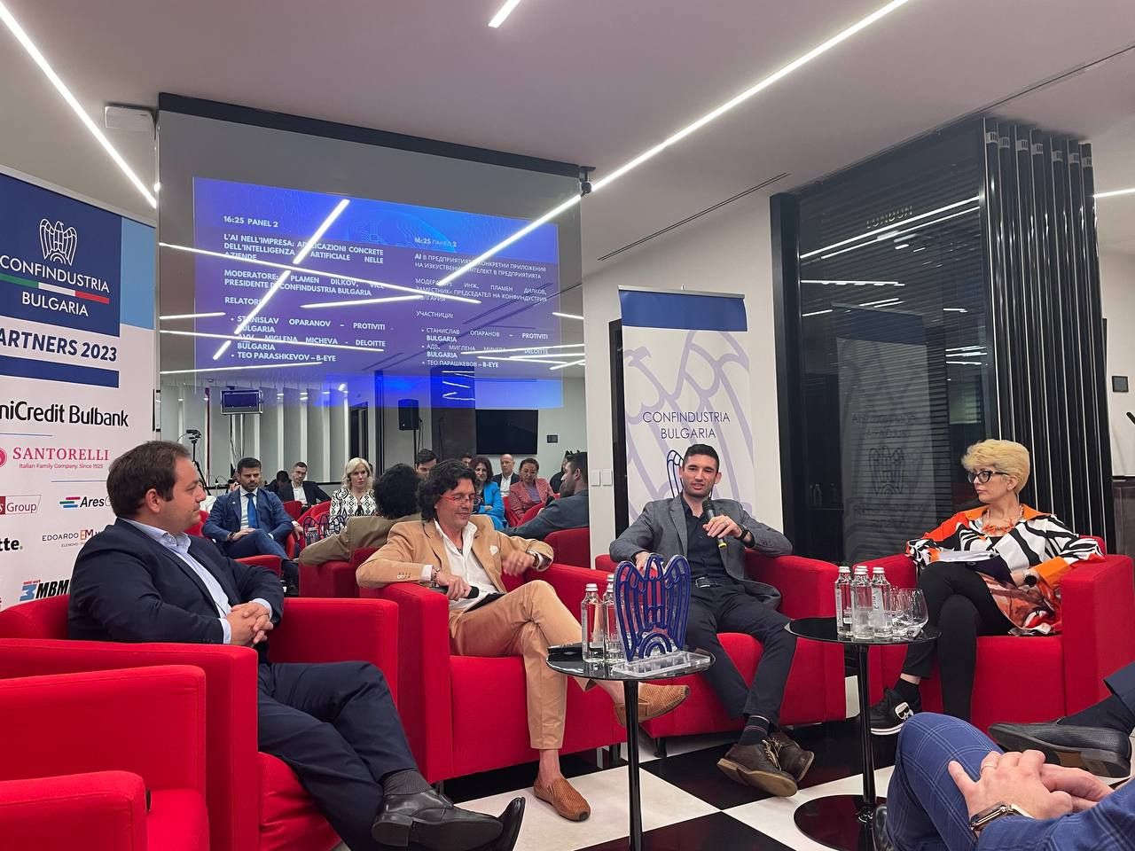 Teo Parashkevov speaking into a microphone during the Enterprise AI Application panel, seated among the other panelists in front of a screen listing the panel participants