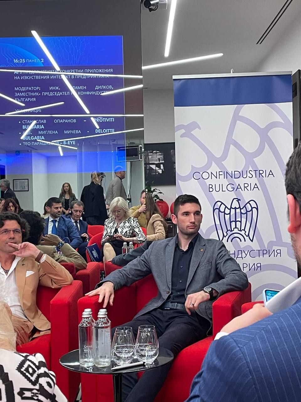 Teo Parashkevov seated in a red armchair on the panel, listening to the discussion beside a Confindustria Bulgaria banner, with the audience in the background
